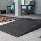 A black rubber threshold ramp on a paved walkway leading to a house entrance with two chairs and plants nearby.