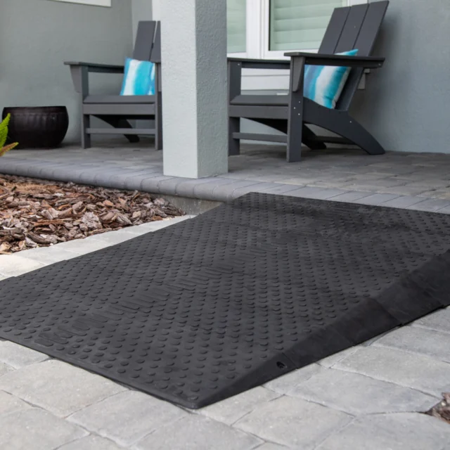 A black rubber threshold ramp on a paved walkway leading to a house entrance with two chairs and plants nearby.
