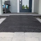A textured black rubber mat on a light gray stone walkway leading to a blue door with a window. A wooden chair is on the left.
