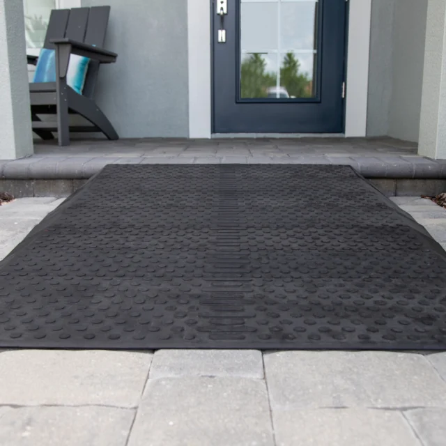 A textured black rubber mat on a light gray stone walkway leading to a blue door with a window. A wooden chair is on the left.