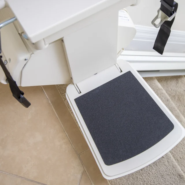 Stair lift platform with straps, positioned at the bottom of a staircase next to a tiled floor.