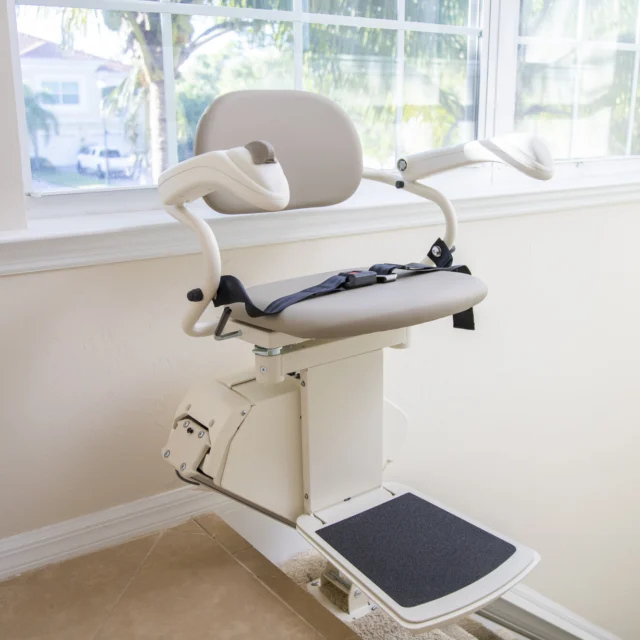 A stair lift with a cushioned seat, armrests, and a safety belt is positioned by a window on a staircase.