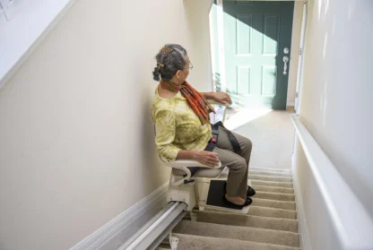 A person using a stair lift chair to ascend a carpeted staircase, facing away from the camera. The stair lift is positioned on the left side of the stairs.