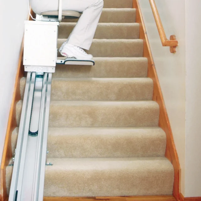 A woman in a red shirt and light pants is sitting on a stairlift chair, positioned on a carpeted staircase with a wooden handrail.