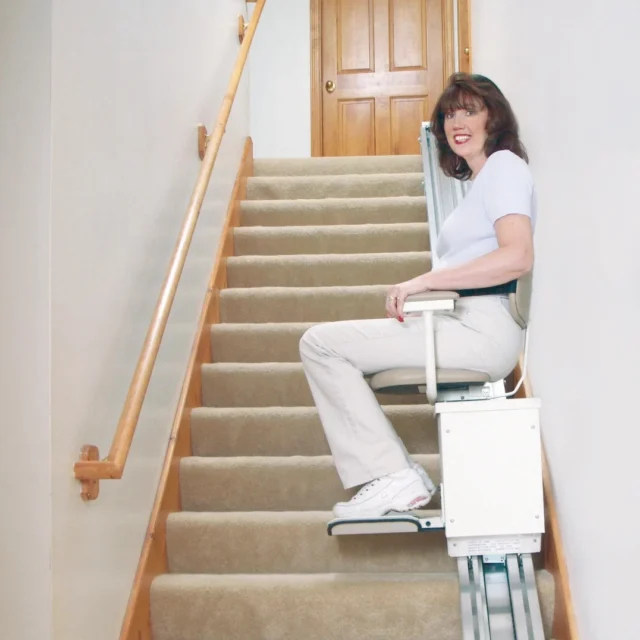 A person sitting on a stairlift at the base of a carpeted staircase, with a wooden handrail on the left and a closed door at the top.