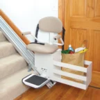 A stair lift with a beige seat is installed on a carpeted staircase. A small platform beside it holds a grocery bag with various items.