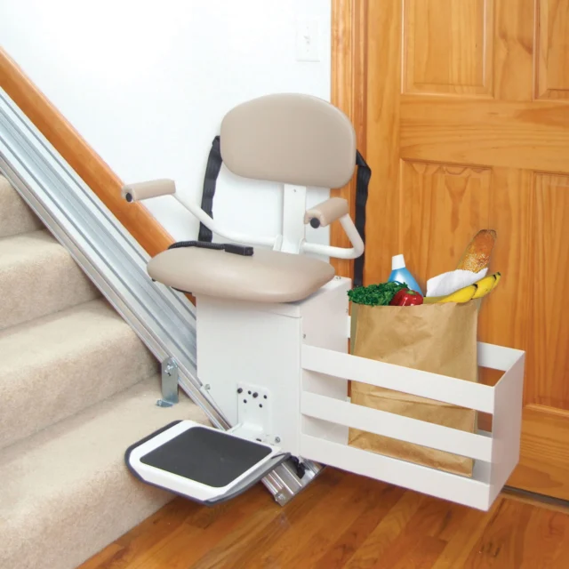 A stair lift with a beige seat is installed on a carpeted staircase. A small platform beside it holds a grocery bag with various items.
