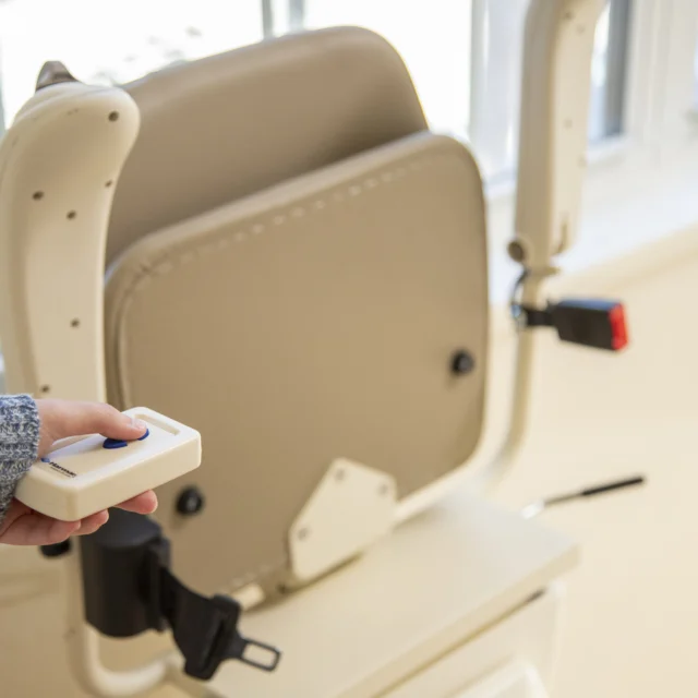 Close-up of a person holding a remote control for a beige stair lift, with its seat and safety belt visible near a window.