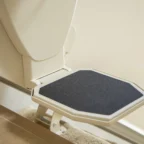 Close-up of a white stair lift footrest with a black non-slip surface on a metal rail, installed over carpet and tile flooring.