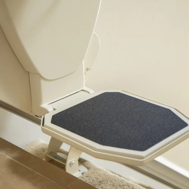 Close-up of a white stair lift footrest with a black non-slip surface on a metal rail, installed over carpet and tile flooring.
