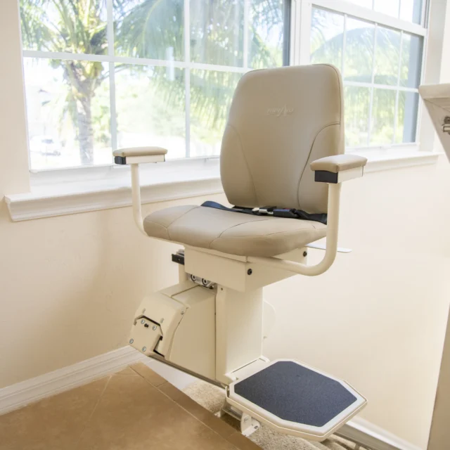 A beige stairlift chair installed next to a staircase, with natural light coming through a large window in the background.
