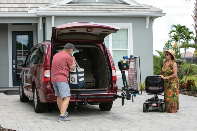 A man loads a bag into the back of a red minivan parked in a driveway. A woman stands nearby beside a mobility scooter on a lift.