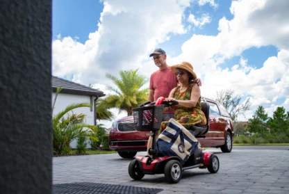 An elderly woman on a mobility scooter and a man walking beside her are outdoors on a sunny day near a home and a parked car.