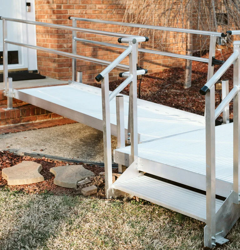A metal wheelchair ramp with handrails leads to a building entrance, set against a brick exterior and surrounded by a lawn and stepping stones.