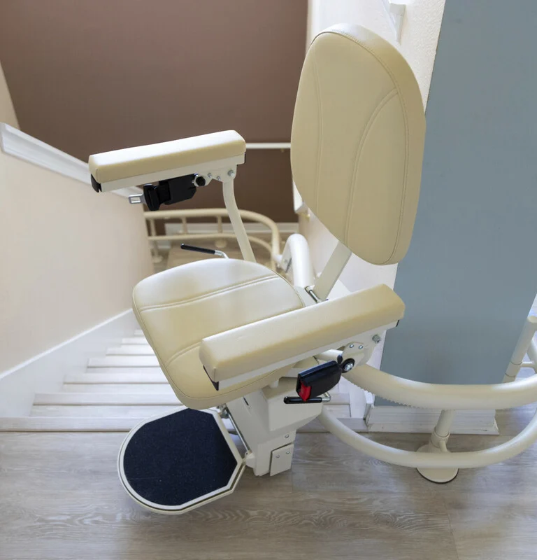 A beige stair lift is installed along a staircase, featuring a padded seat and backrest with armrests, ready for use.