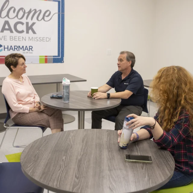 Three people sit at round tables in a room with a "Welcome Back" banner. Two women and one man are having a conversation. A water bottle and a smartphone are on the table.