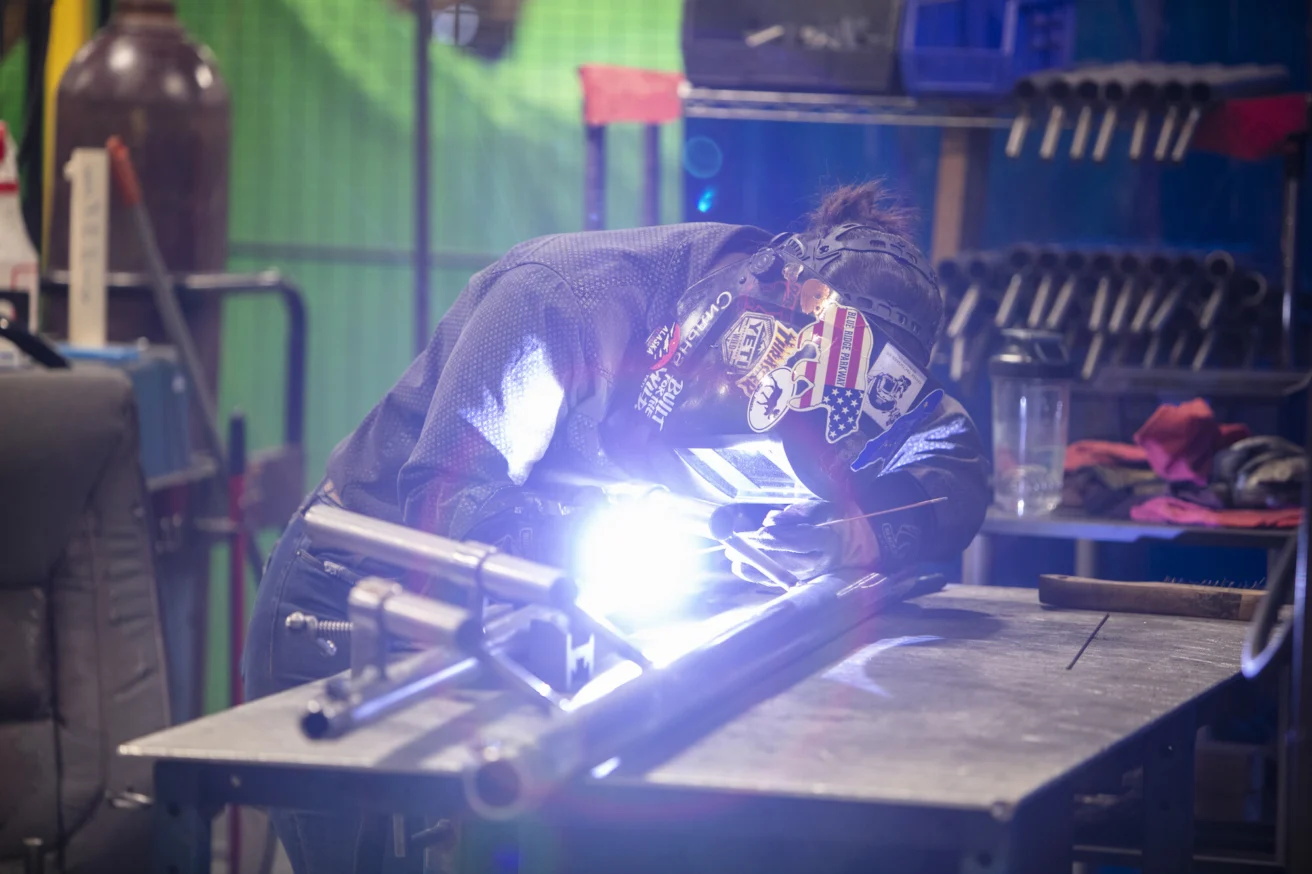 A person welding metal pipes on a table wears a protective helmet with various stickers. Bright sparks and a blue light emanate from the welding process in an industrial setting.