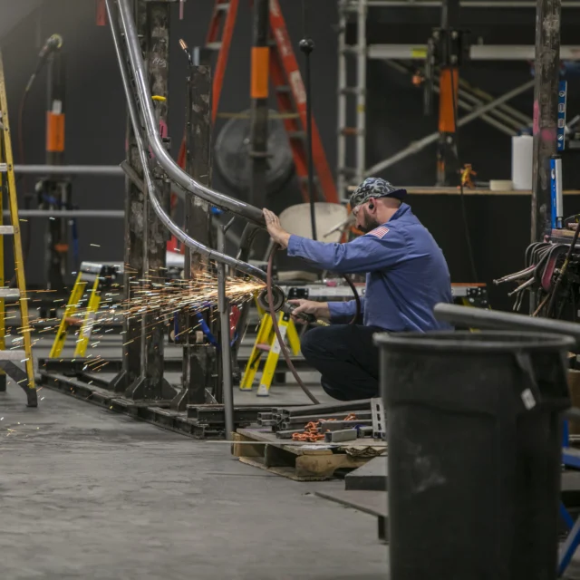 A person wearing a headscarf operates a tool, grinding metal and creating sparks in an industrial workshop with ladders and various equipment around.