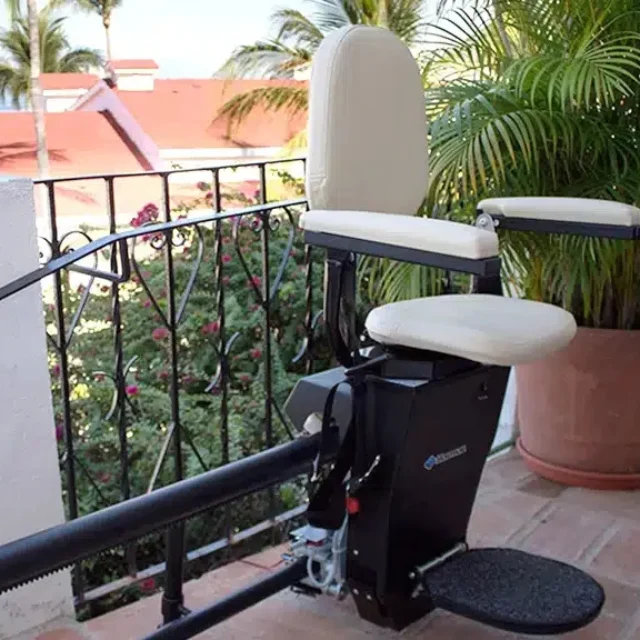 A stairlift installed on an outdoor staircase with a decorative railing, set against a backdrop of red rooftops, trees, and a view of a distant body of water.