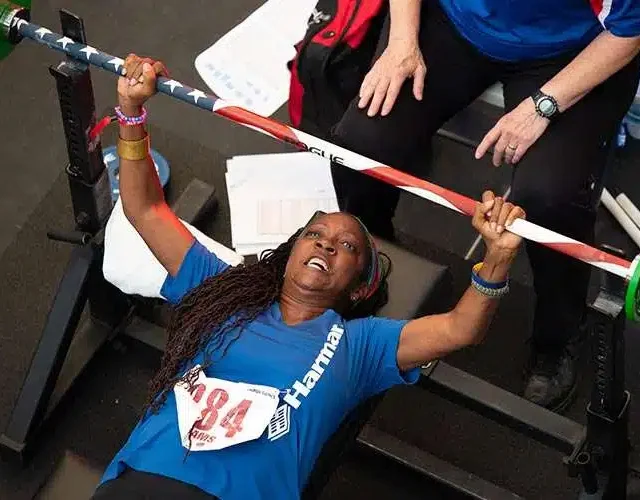 A person in a blue shirt is performing a bench press with a barbell covered in a flag-themed pattern. An instructor in a blue shirt observes nearby.