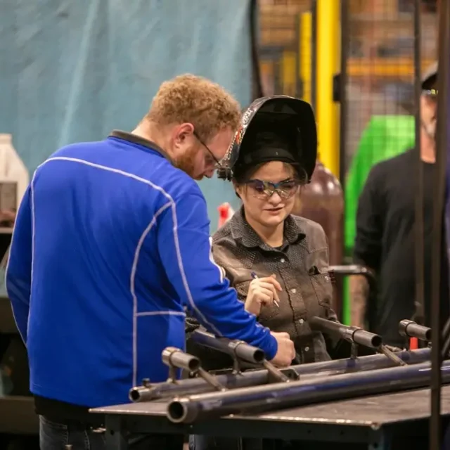 Two people in a workshop review metal tubing. One person wears a welding helmet, and the other wears a blue jacket.