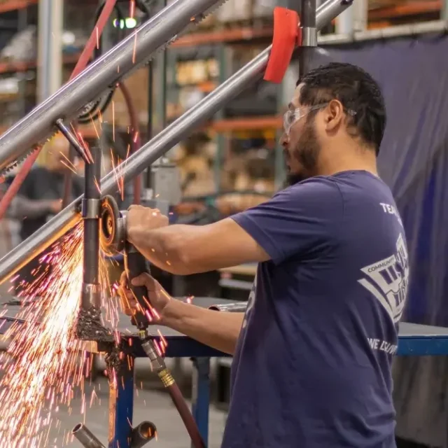 A man wearing safety goggles uses a grinder on a metal frame in a workshop, creating sparks. Shelves with materials are visible in the background.