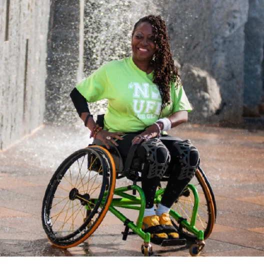 Person in a bright green T-shirt and adaptive wheelchair smiling near a fountain.