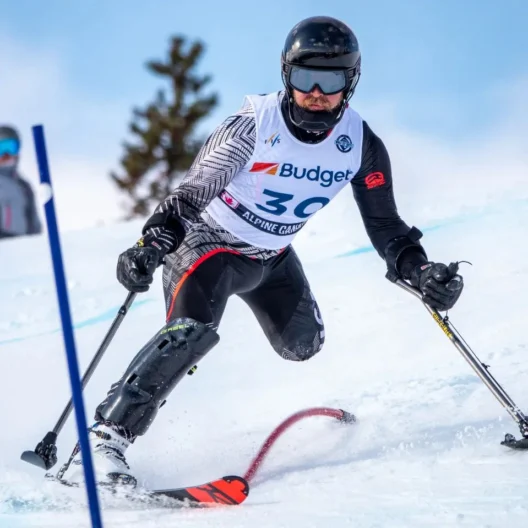 A skier competes in a slalom event, wearing a helmet and adaptive equipment on a snowy slope, with others watching in the background.