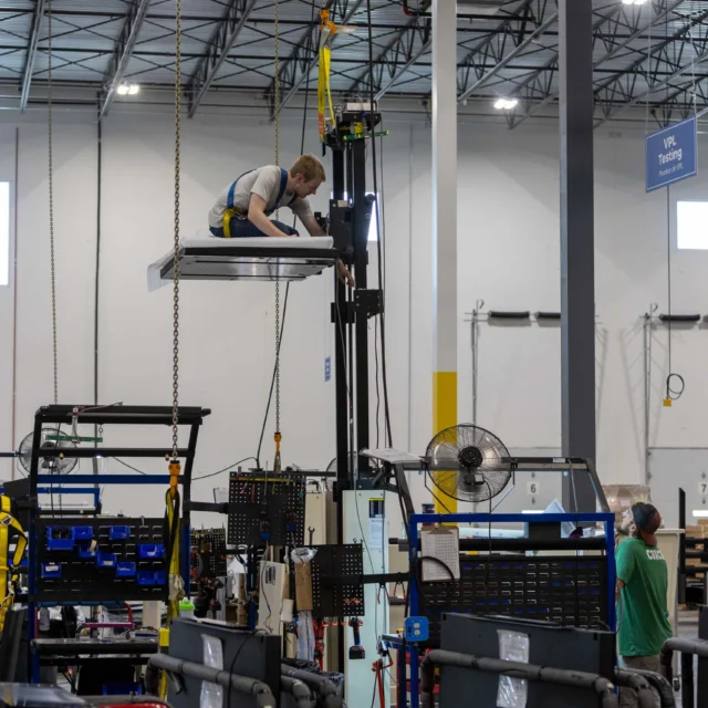 Worker in a harness perched on elevated platform inside an industrial facility with another person standing below, surrounded by machinery and equipment.