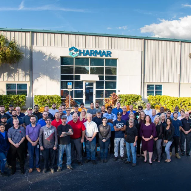 A large group of people stands in front of a building with "Harmar" signage. The scene is outdoors on a clear day.