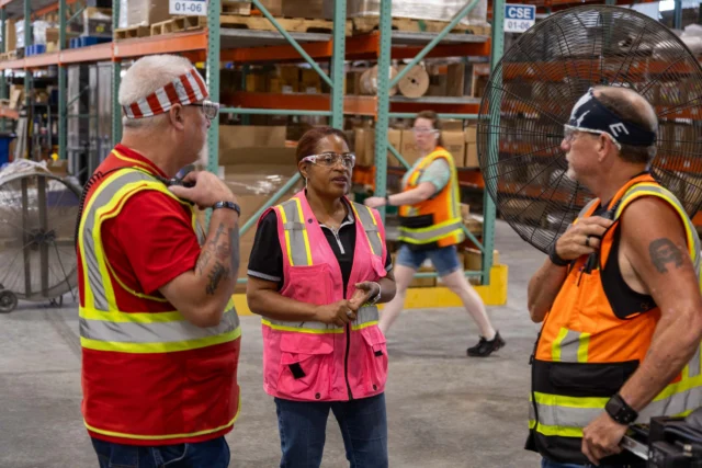 Three people wearing safety vests and glasses stand talking in a warehouse, while another person walks by holding a clipboard. A large industrial fan is visible in the background.