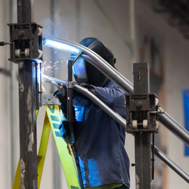 A person in protective gear is welding metal pipes on a construction site. Sparks are visible, and a yellow ladder is nearby.