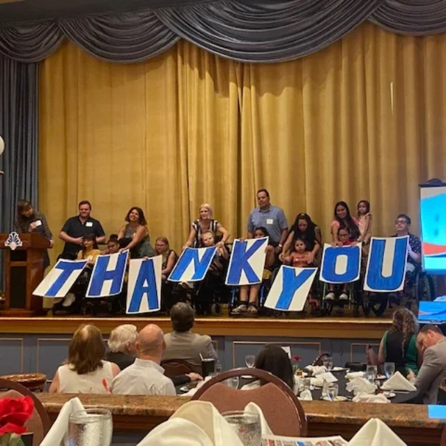 People holding large letters spelling "THANK YOU" on stage at the 13th Annual Heroes Luncheon, with attendees seated at tables.