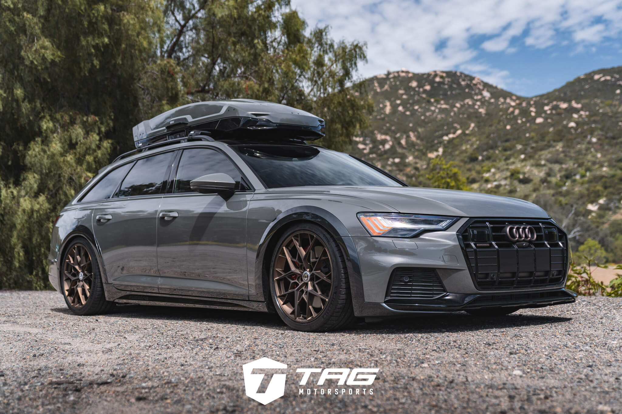 A sleek gray Audi station wagon with a roof box, featuring bronze wheels, parked on a gravel road with a mountainous backdrop.
