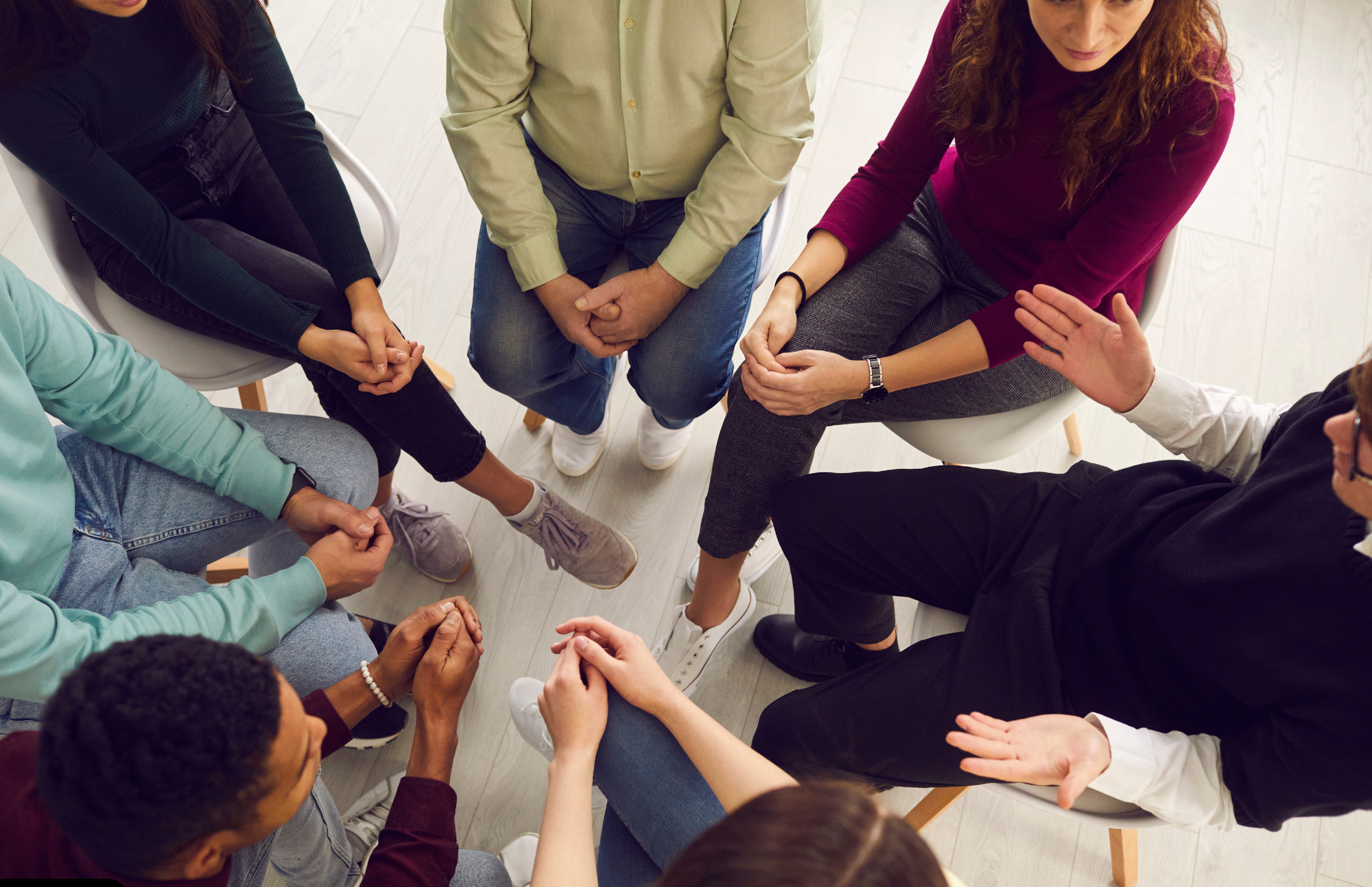 A group of people sits in a circle, engaged in a discussion at AMFM Healthcare. The photo is taken from above, highlighting their hands and legs as they gesture and listen attentively to each other.