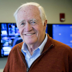 An older man with white hair smiles at the camera, wearing a rust-colored sweater over a blue collared shirt. He stands in front of large monitors displaying blue screens in an office setting.