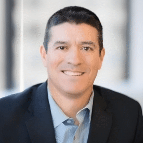 A man with short dark hair wearing a dark blazer and light blue shirt smiles at the camera in a bright, blurred office setting.