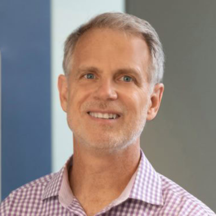 A middle-aged man with short gray hair and a trimmed beard smiles at the camera. He is wearing a light purple checkered shirt, representing Lineage Capital, a private equity investment firm. The background is blurred with blue and gray tones.