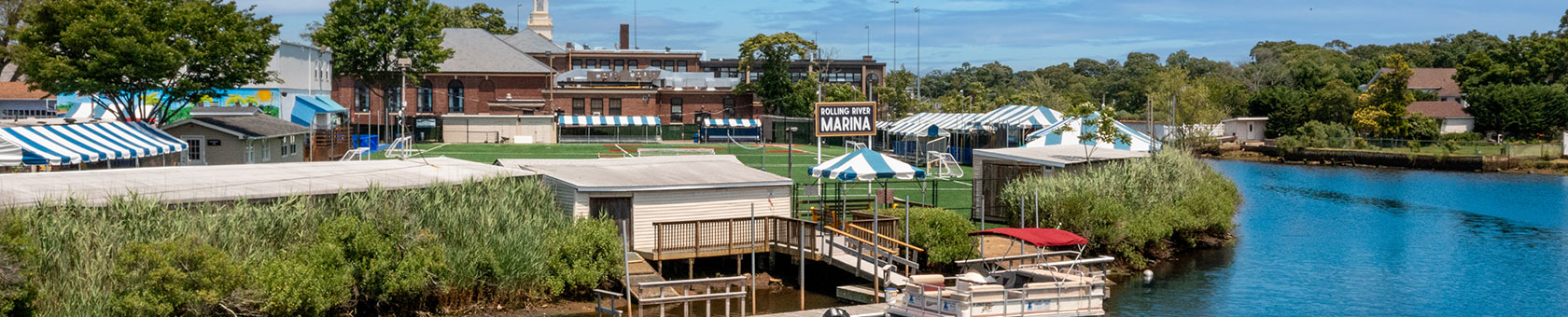 View of a small marina with striped tents, docks, and boats along a calm waterway, with buildings and trees in the background under a blue sky.