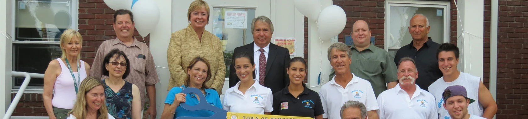 A group of people poses outdoors in front of a brick building, some holding a large yellow sign. White balloons decorate the area.