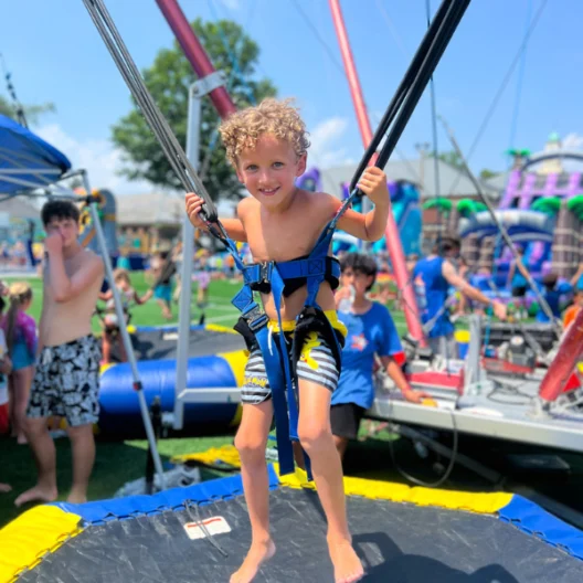 Young boy wearing a harness stands on a trampoline at an outdoor fair, with other children and inflatable structures in the background.