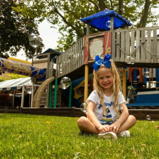 Young girl with a blue bow sits on grass in front of a playground, smiling, with bubbles floating around her on a sunny day.
