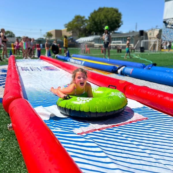 A young girl slides down an outdoor slip-and-slide on a green inflatable tube while other children wait in line on a sunny day.