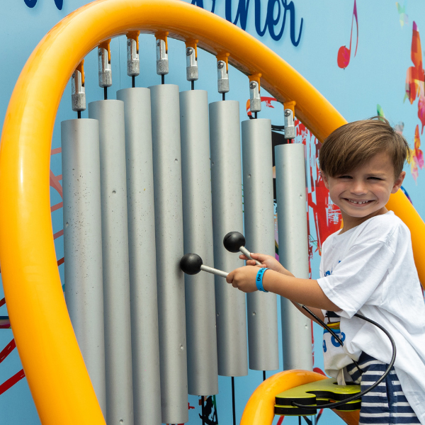 A young boy smiling while playing large outdoor chimes with two mallets, standing in front of a colorful wall.