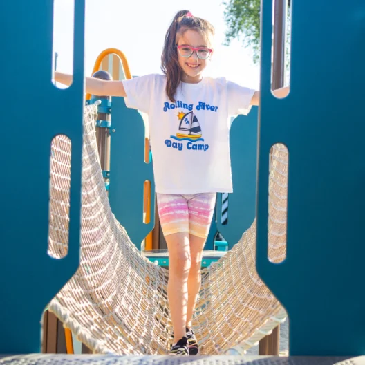 A young girl wearing glasses and a "Rolling River Day Camp" T-shirt stands on a rope bridge at a playground, smiling at the camera.