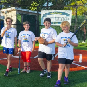 Four children in matching "Rolling River Day Camp" shirts stand on a baseball field, smiling and holding baseball equipment.