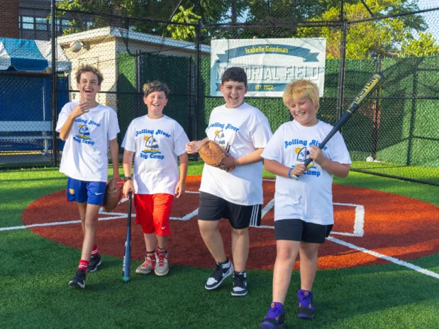 Four children in matching "Rolling River Day Camp" shirts stand on a baseball field, smiling and holding baseball equipment.