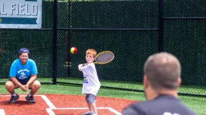 A young boy prepares to hit a baseball with a racket while a coach pitches and another person crouches behind him on an outdoor field.