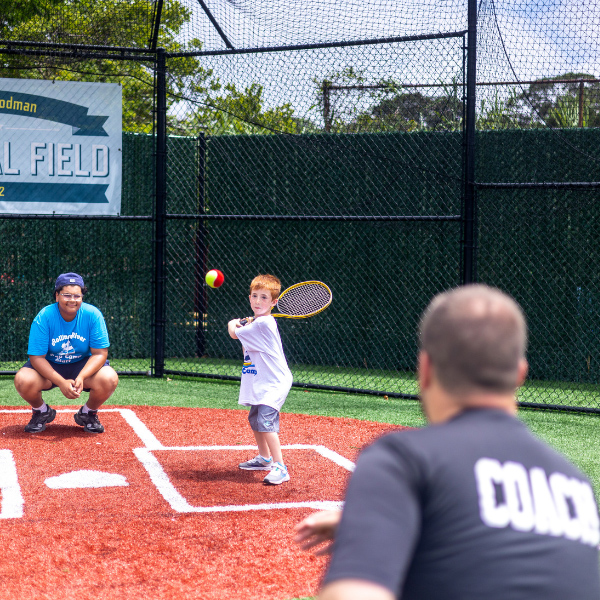 A young boy prepares to hit a baseball with a racket while a coach pitches and another person crouches behind him on an outdoor field.