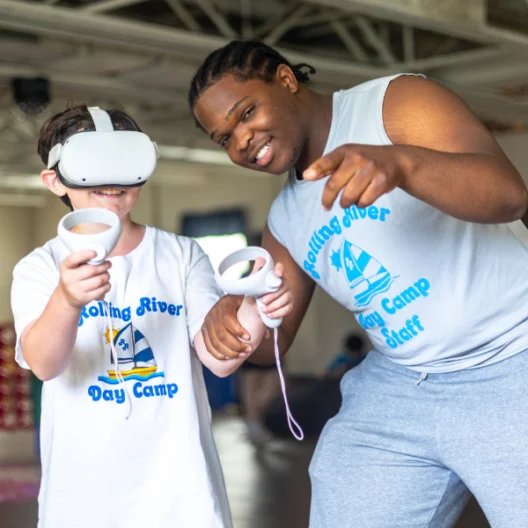 A camp counselor helps a child wearing a VR headset and holding controllers at Rolling River Day Camp. Both are smiling and wearing matching camp shirts.